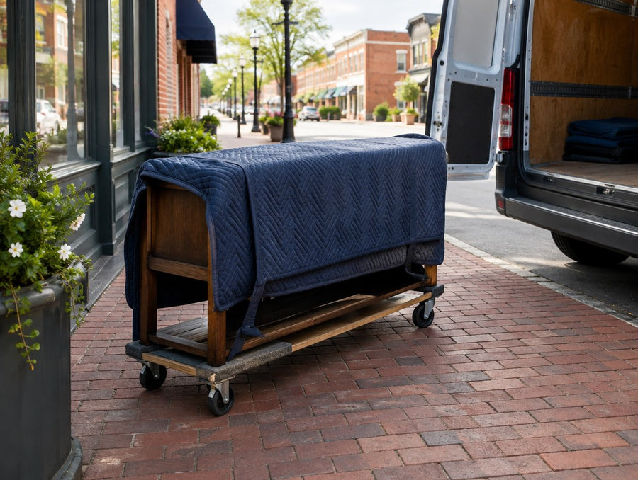 A blanket-wrapped solid wood console on a dolly beside a delivery van on a brick main street